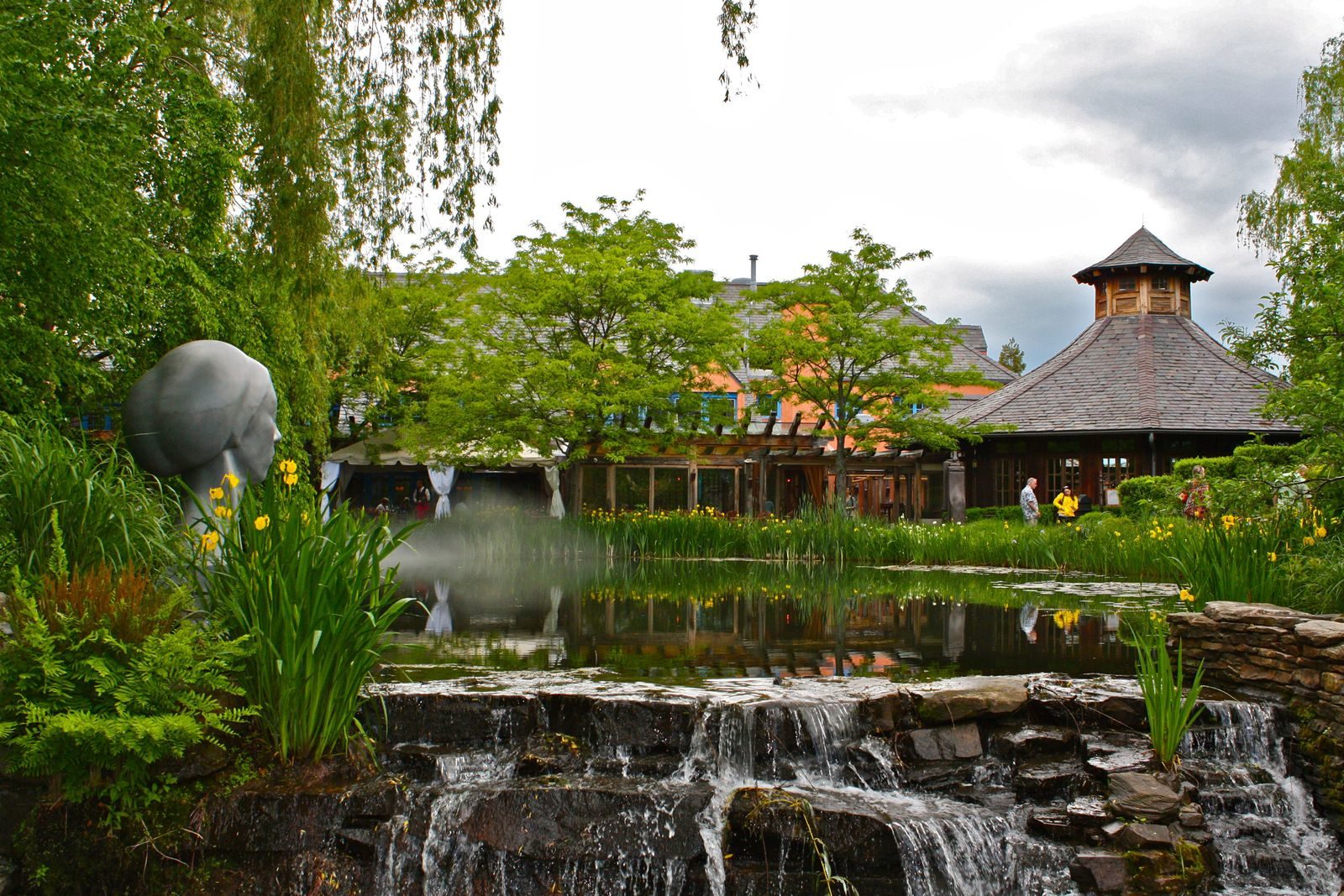 Scenic garden with a pond, waterfall, and sculpture under a cloudy sky.